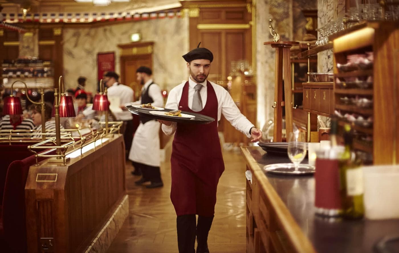 A waiter carrying a tray through a grand art deco dining room